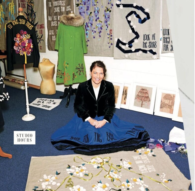  Kate wearing a black velvet jacket, white v neck t-shirt, and long blue skirt, is sitting on the floor of her studio wearing a long blue skirt surrounded by some of her art pieces.  Directly in front of her is a large piece with lots of white and yellow flowers and the words 