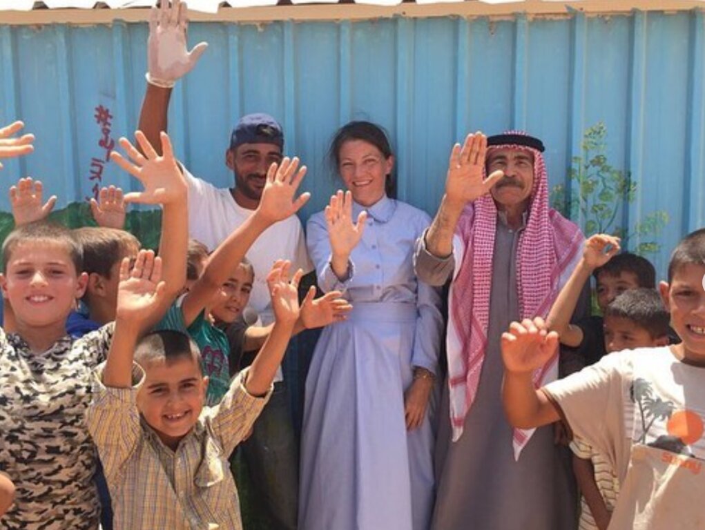 A Photo of Kate standing against a shipping container surrounded by middle eastern refugees two adult men and lots of children everyone is smiling and waving at the camera