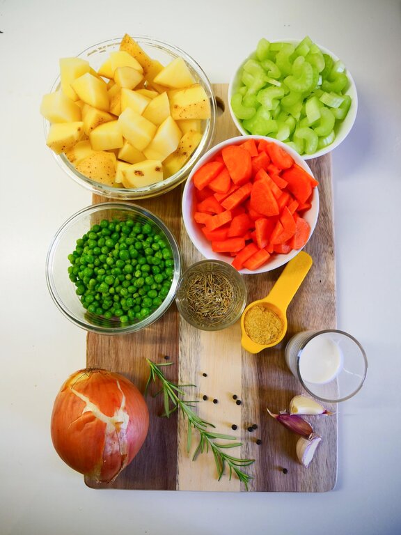  Alt Text: A wooden chopping board on a white surface on the board are the ingredients peas, chopped carrots, Celery sticks and chopped potatoes in separate bowls and herbs and spices on the board