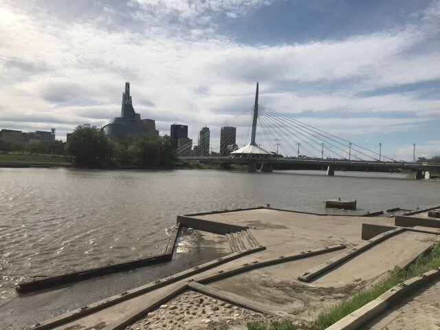  Alt Text: A view of the Red River riverside walkway at St Boniface in Winnipeg looking towards the Canadian Museum of Human Rights on the left and the Esplanade Reil Bridge on the right.  In the background are the Winnipeg business district high-rise offices.